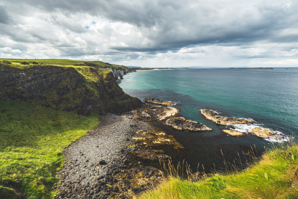 Irish shoreline under the cloudy sky background. Northern Ireland. Green covered land surrounded by the water surface. Picturesque Irish landscape. Perfect place for the outdoor activity.