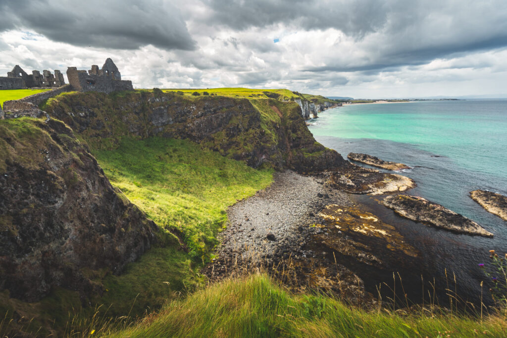 Northern Ireland shoreline with Dunluce castle in the distance. Picturesque grass covered lowland and the steep cliffs under the grey cloudy sky. Amazing Irish bay before the rain. Wild virgin nature.