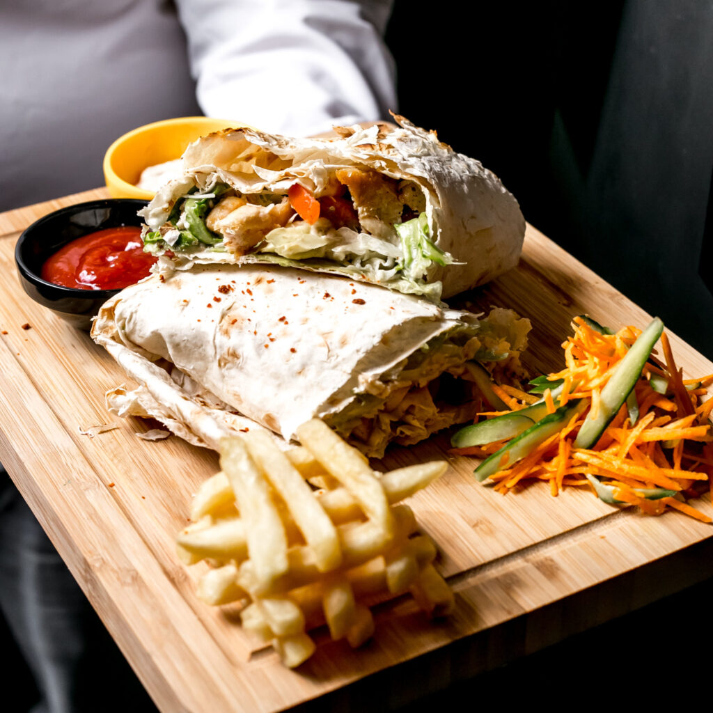 side view a man holds a tray with chicken doner in pita bread with ketchup mayonnaise french fries and vegetable salad on the board