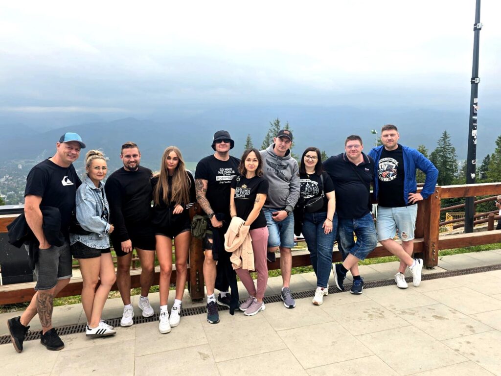 Group of people posing together on a mountain viewpoint terrace with a wooden railing, overlooking a distant valley under a cloudy sky.