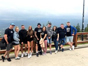 Group of people posing together on a mountain viewpoint terrace with a wooden railing, overlooking a distant valley under a cloudy sky.