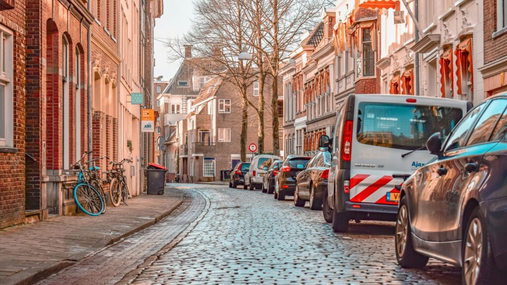 A narrow, cobblestone Dutch street lined with brick row houses, dozens of parked cars and a few bicycles squeezed against the sidewalk—illustrating one of the “bad things about living in Netherlands”: crowded parking and tight urban layouts.