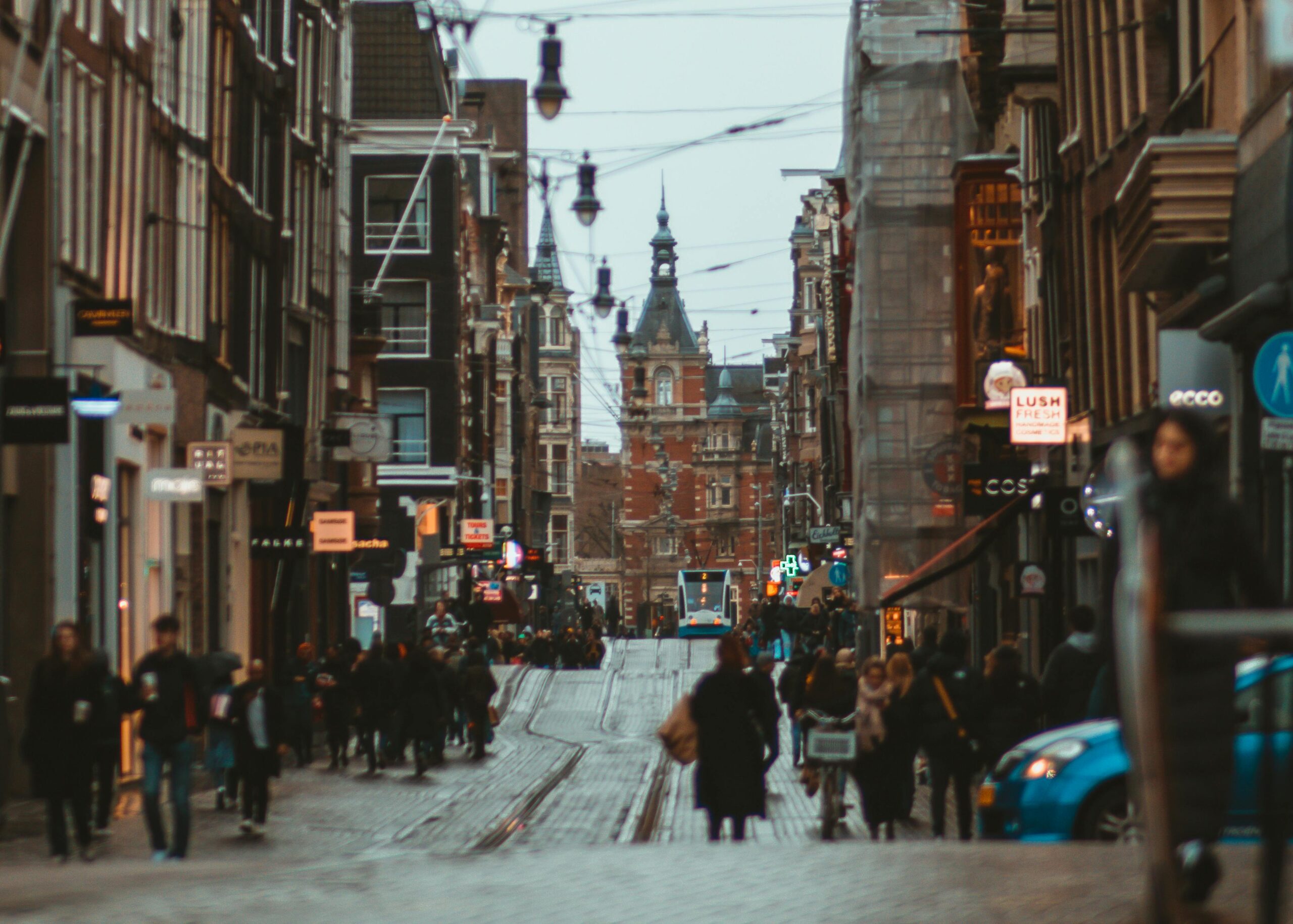 A busy Amsterdam shopping street with historic façades, overhead tram wires, and a tram approaching in the distance, as locals and expats in Netherlands stroll and cycle along the wet cobblestones.