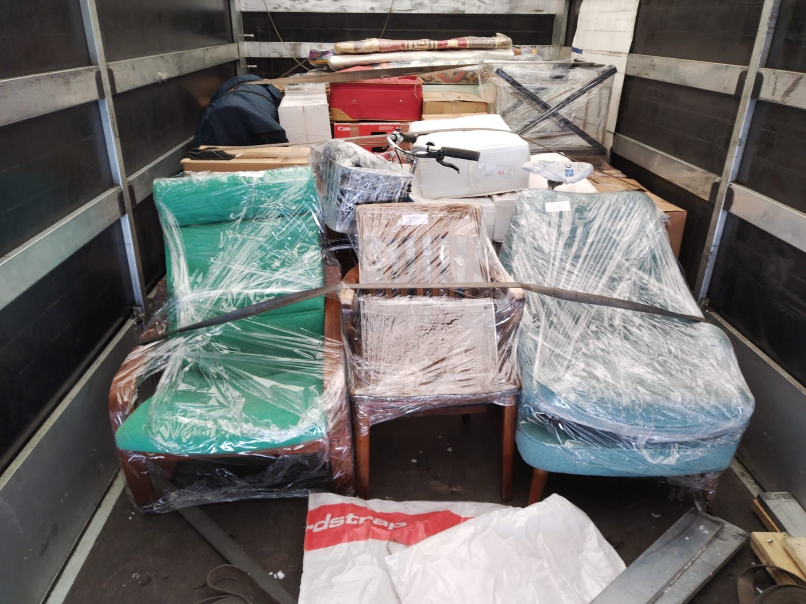Inside a moving van loaded for an international relocation: three armchairs wrapped in clear plastic film at the front, with stacked cardboard boxes, a wrapped bicycle basket, rolled-up rugs, and other household items secured behind them.
