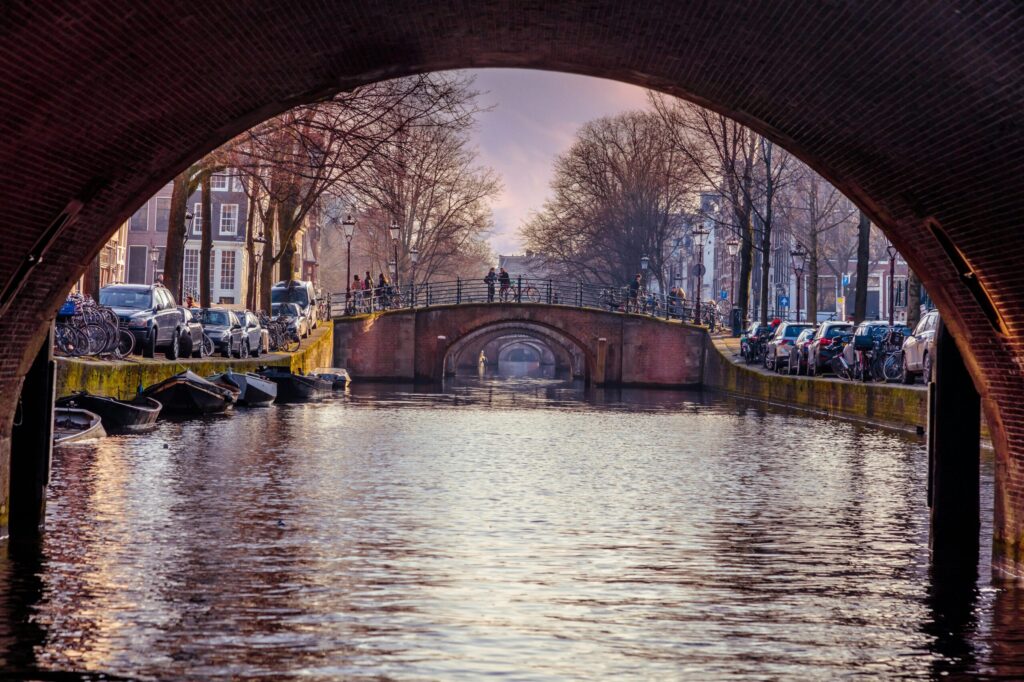View under a canal bridge in Amsterdam at sunset with iconic houses and moored boats – capturing the charm and raising the thought: how expensive is Amsterdam?