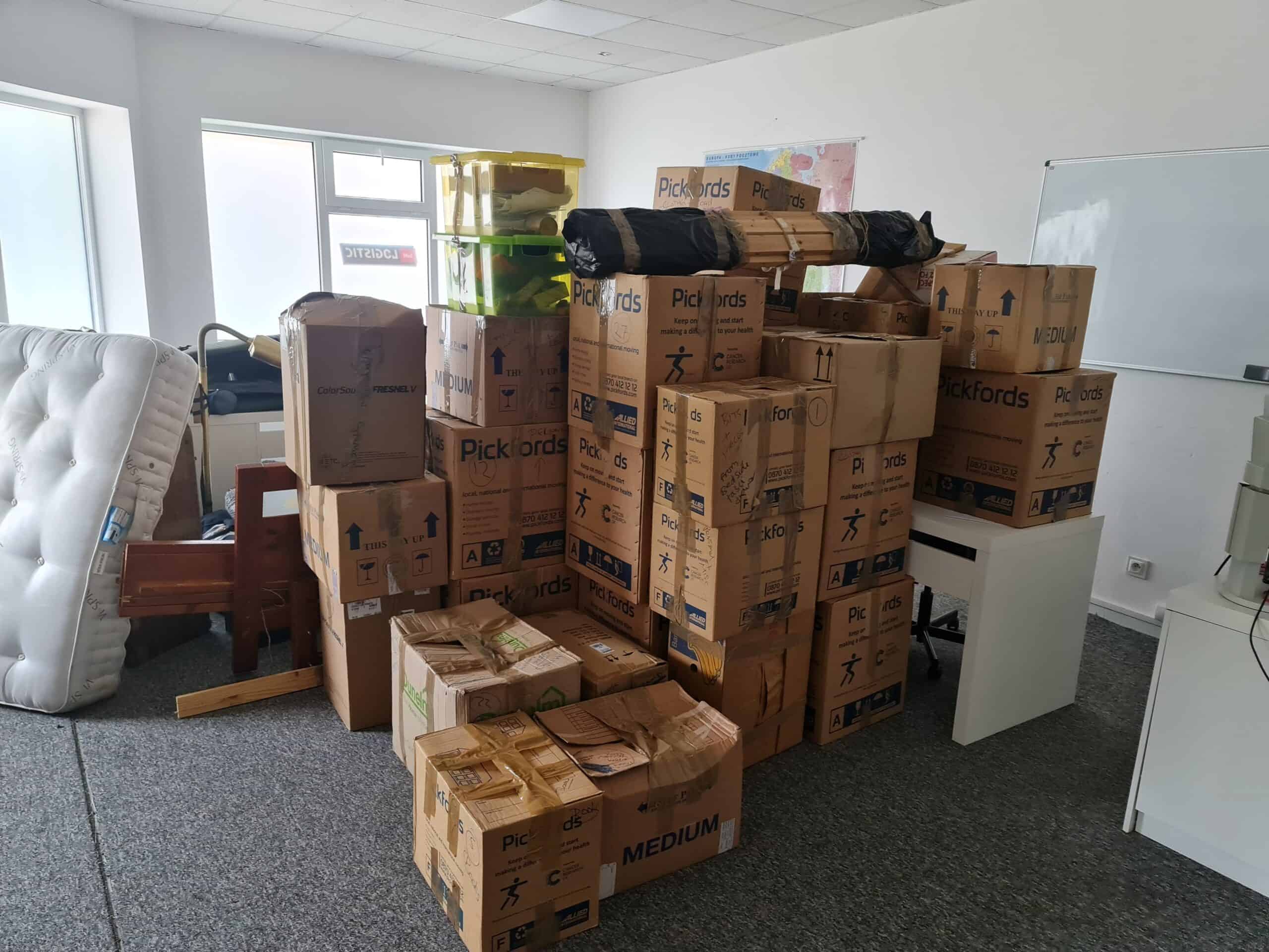 Room filled with stacked moving boxes, furniture, and packed items, all organized for moving from UK to Amsterdam.