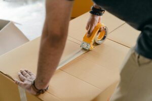 Hands sealing a large cardboard box with packing tape, preparing items for a move to the Netherlands from the UK.
