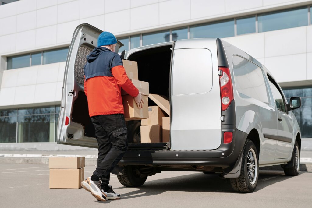 A mover in a blue beanie and red jacket lifts cardboard boxes into the back of a silver van parked outside a modern building, illustrating a relocation to the Netherlands.