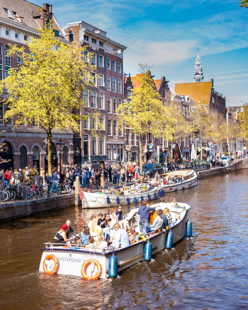 Tour boat cruising an Amsterdam canal past crowded quayside cafés, showcasing one of the first experiences when relocating to the Netherlands.