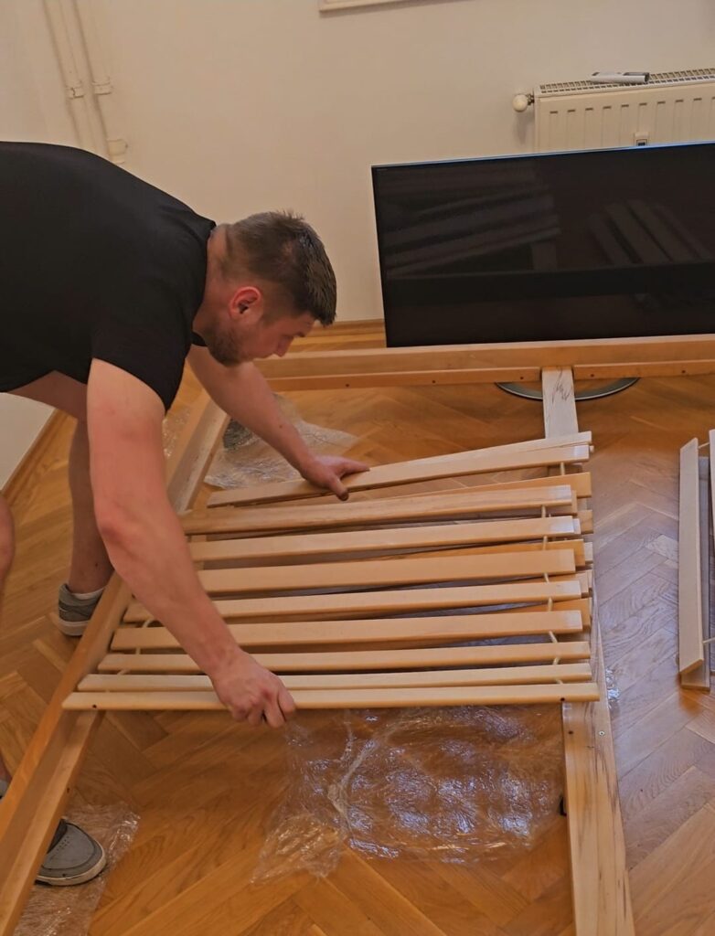A mover disassembles a wooden bed frame on a parquet floor, preparing furniture for a relocation from Berlin to Budapest.