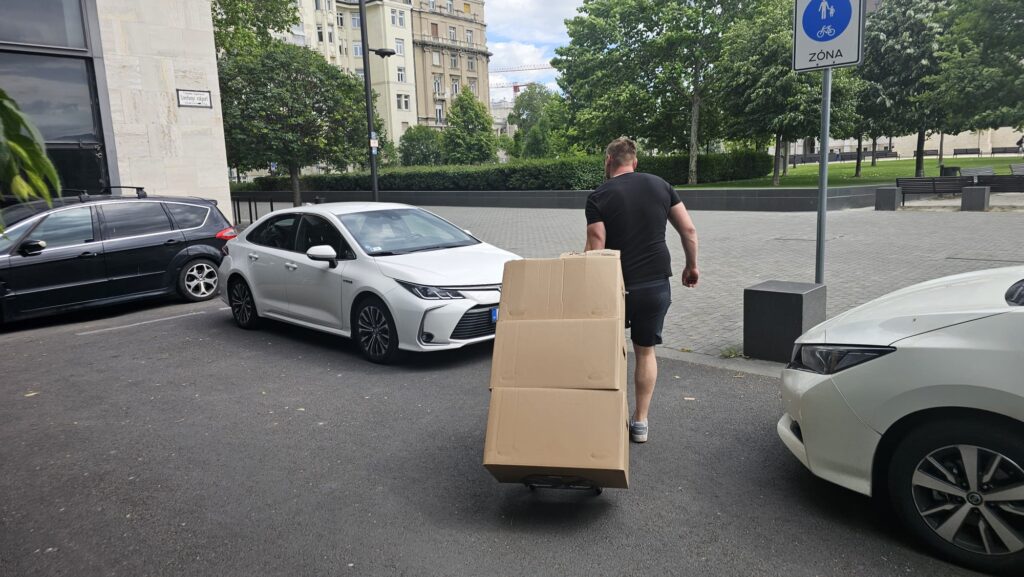 A mover pulls a hand truck loaded with large cardboard boxes along a street during a Berlin to Budapest relocation.