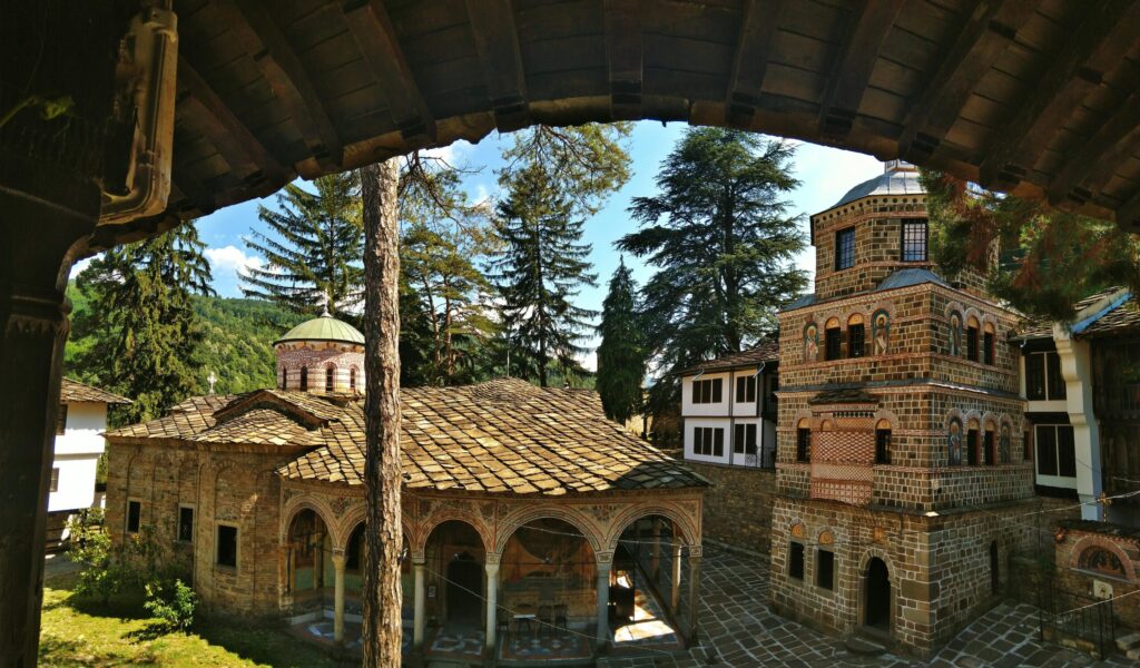 Traditional Bulgarian monastery courtyard with historic stone buildings, frescoed arches, and pine trees in a mountain setting.