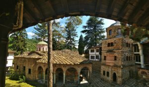 Traditional Bulgarian monastery courtyard with historic stone buildings, frescoed arches, and pine trees in a mountain setting.