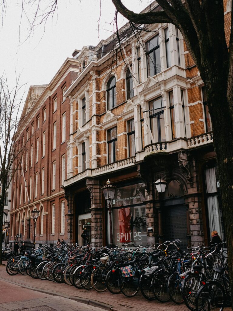 A row of parked bicycles locked to a metal railing along a brick-paved street in front of historic Dutch canal-side buildings.