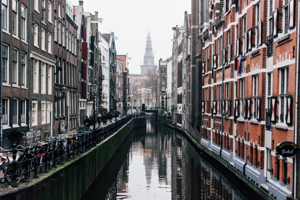 A quiet Amsterdam canal flanked by tall, narrow brick townhouses with shuttered windows and bicycles locked along the railing.