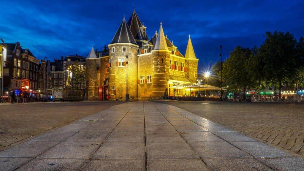 A historic brick gatehouse (De Waag) in Amsterdam’s old town square, lit warmly at dusk with surrounding cafés and tram tracks.