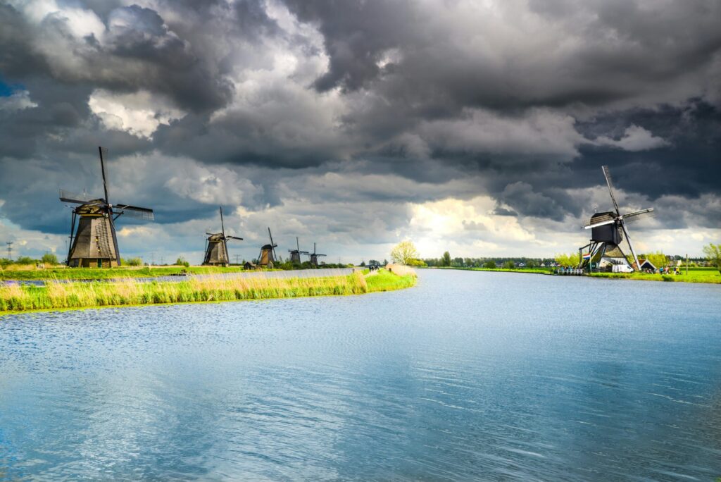 A row of traditional Dutch windmills standing beside a wide canal under a dramatic, cloudy sky, with green grassy banks in the foreground.