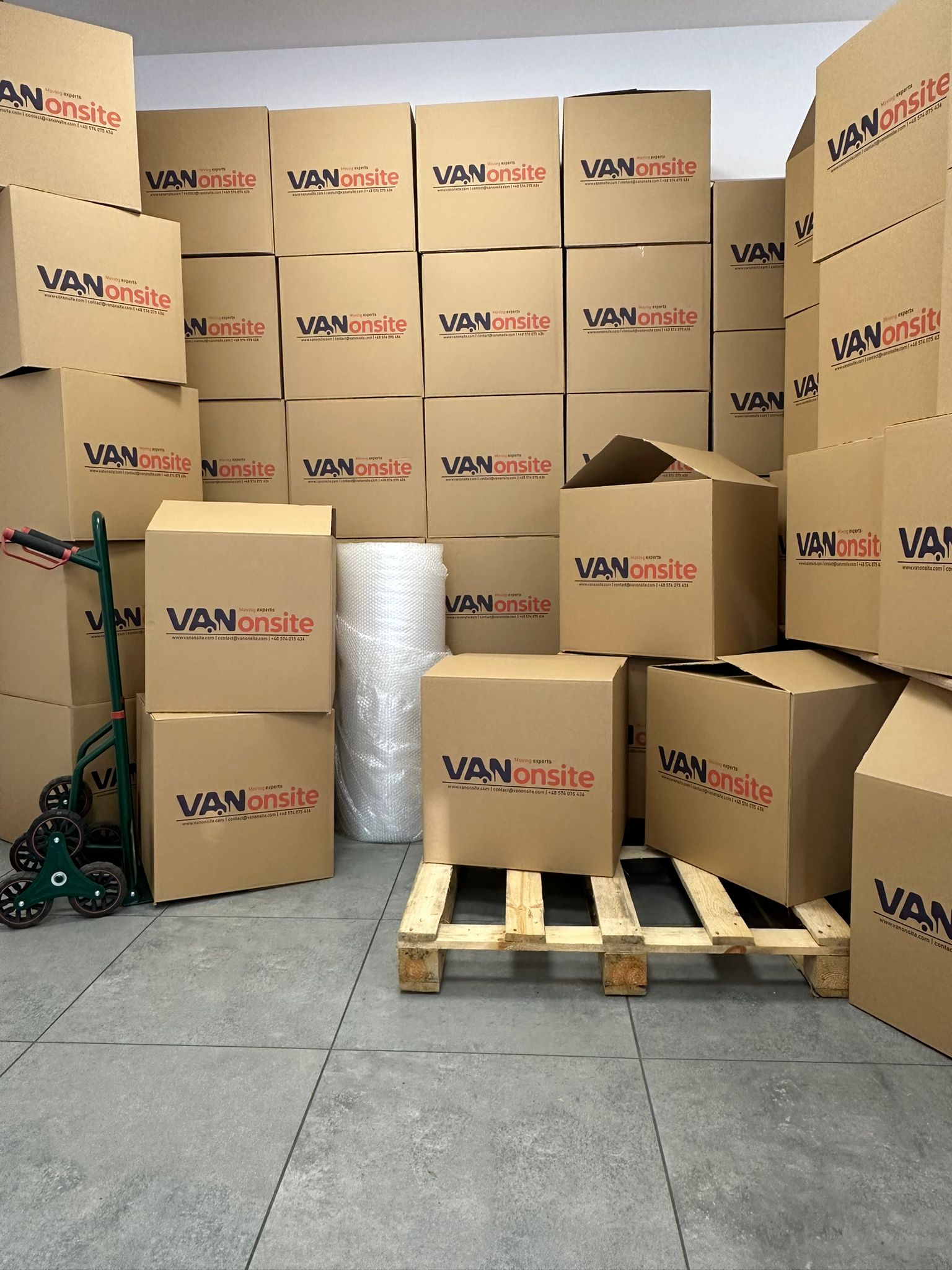 Stacked VANonsite moving boxes in a storage area, with a pallet, hand truck, and packing wrap ready for a move