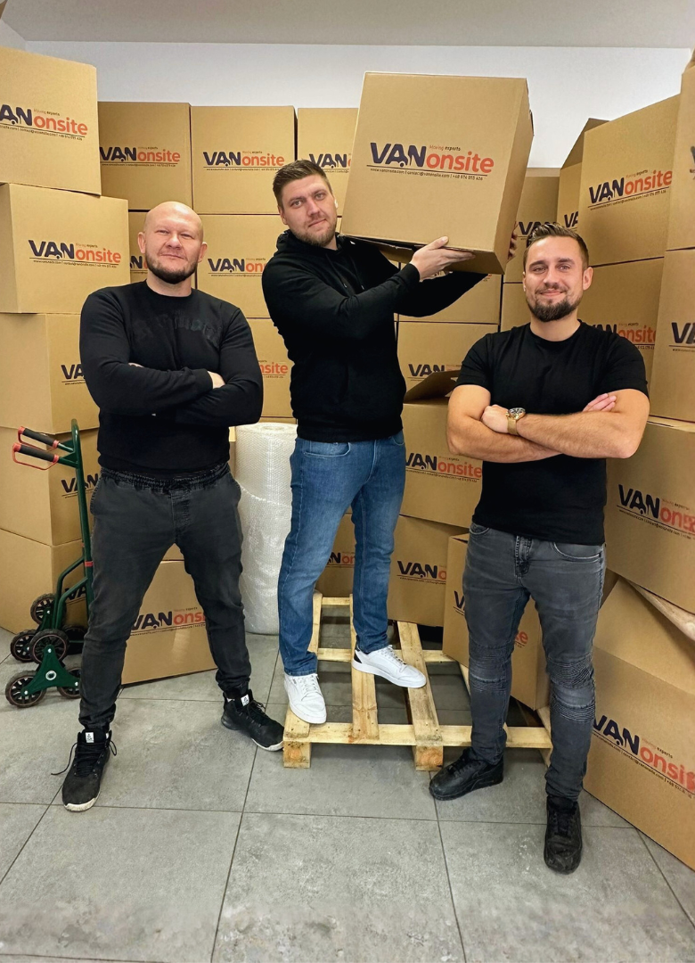 Three VANonsite team members in a warehouse surrounded by branded moving boxes, one holding a box for packing