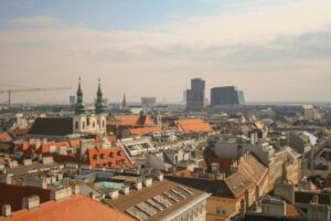 Panoramic city skyline with red rooftops, twin church towers, and modern high rise buildings in the distance under a hazy sky.