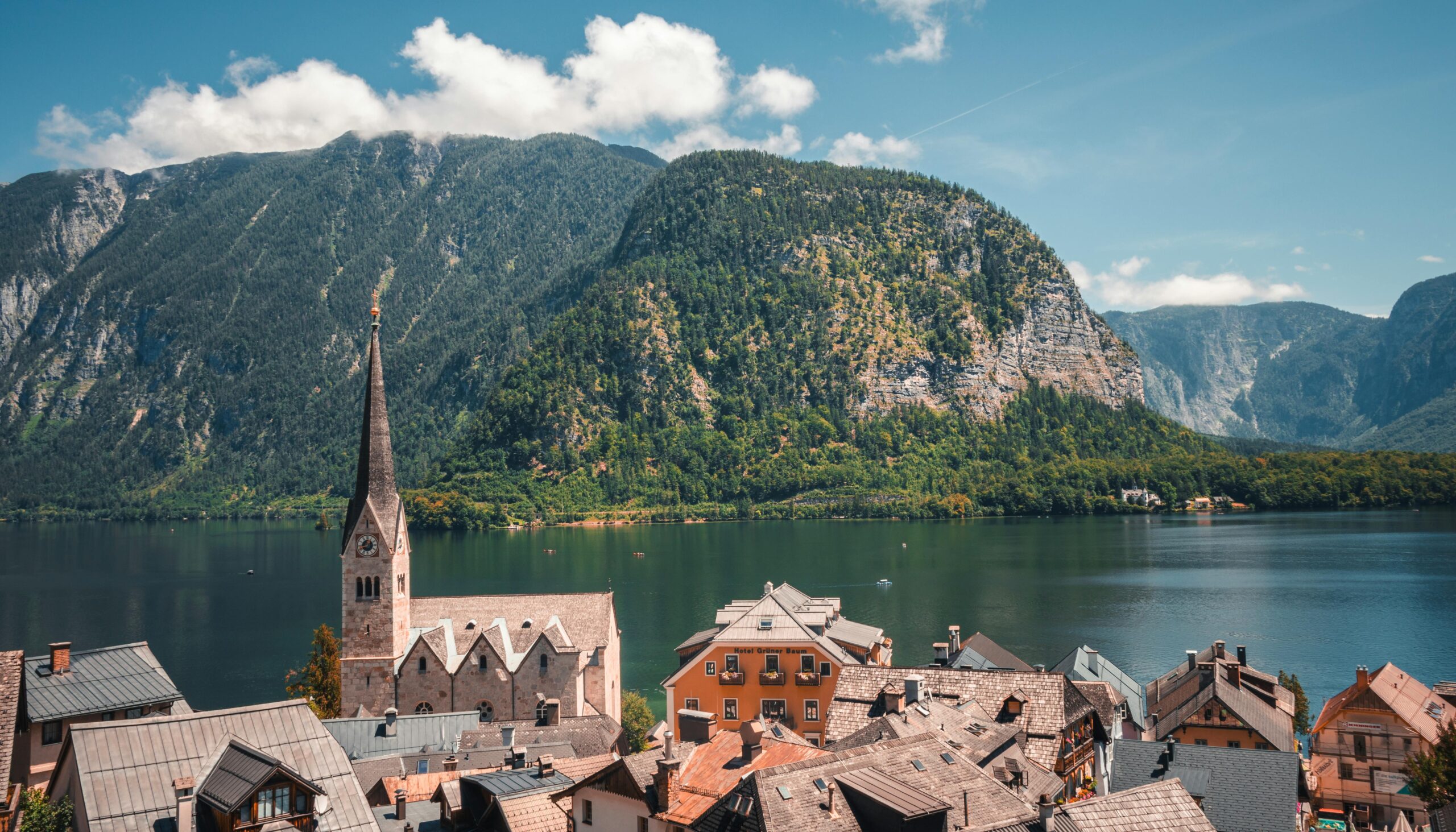 Alpine lakeside village with a tall church steeple, rooftops in the foreground, and forested mountains across a calm lake under a blue sky.