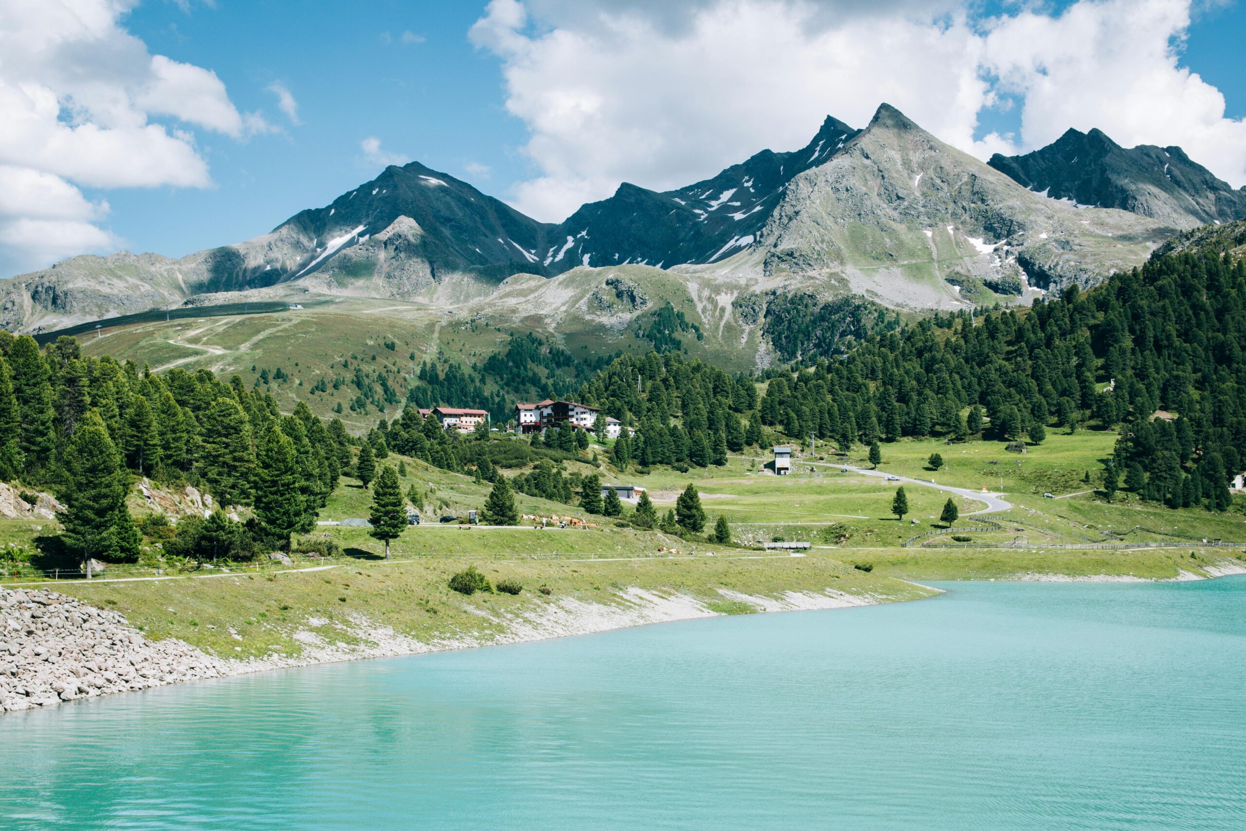 Turquoise alpine lake below rugged mountain peaks, with pine forest and a small lodge on a green hillside under a partly cloudy sky.
