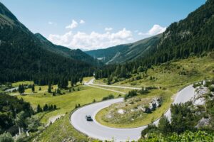 Winding mountain road with a hairpin turn through a green valley, with a car and forested hills under a blue sky.