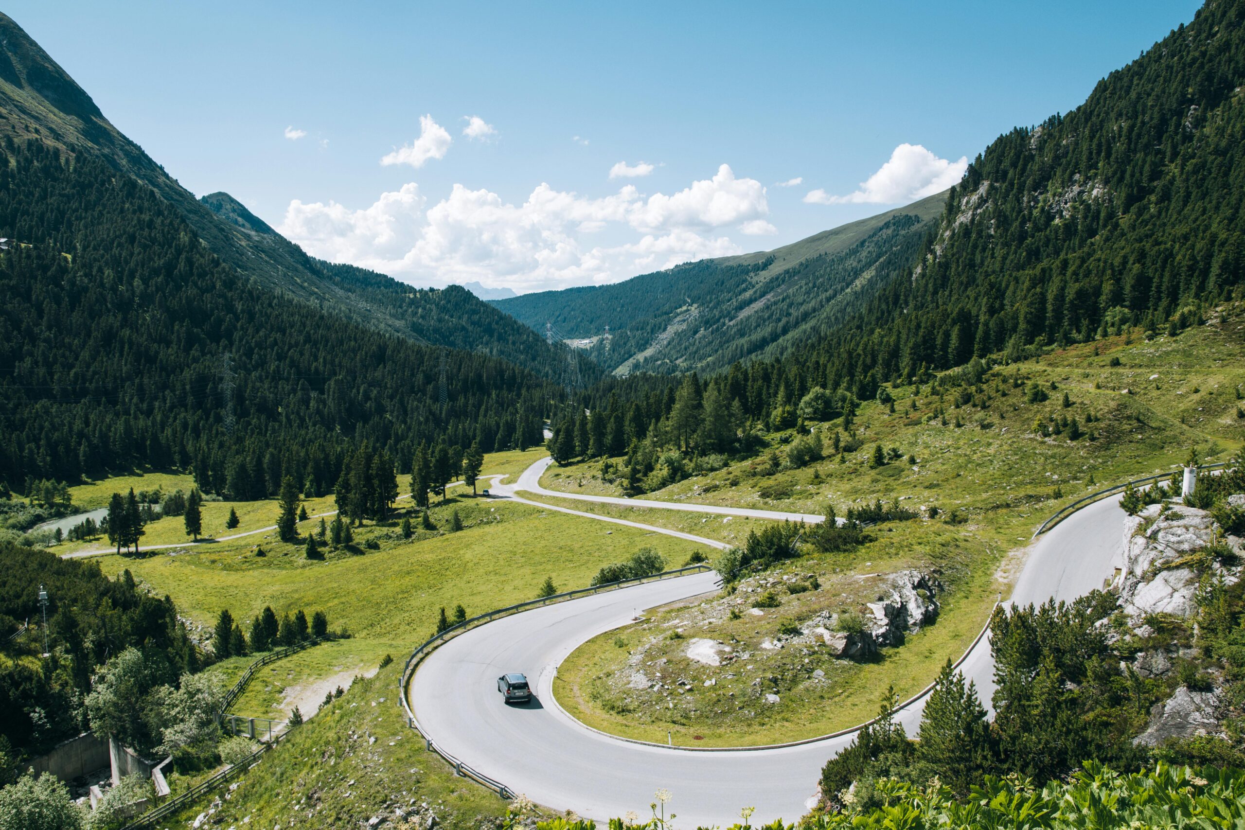 Winding mountain road with a hairpin turn through a green valley, with a car and forested hills under a blue sky.