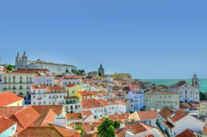 Colourful coastal city skyline with red tiled rooftops, historic churches, and the sea under a clear blue sky