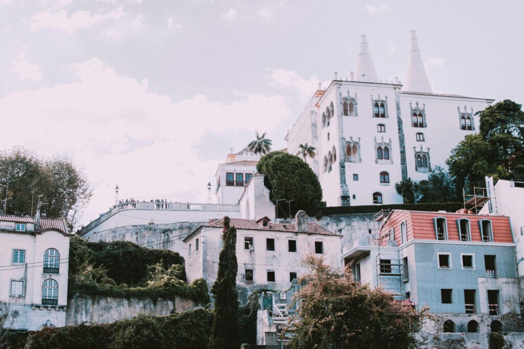 White historic palace on a hillside above colourful houses and greenery under a soft sky