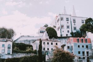 White historic palace on a hillside above colourful houses and greenery under a soft sky