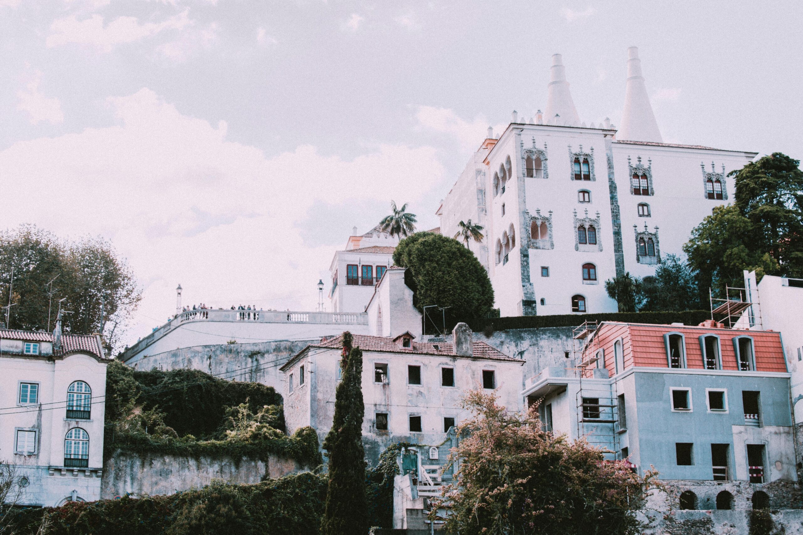 White historic palace on a hillside above colourful houses and greenery under a soft sky