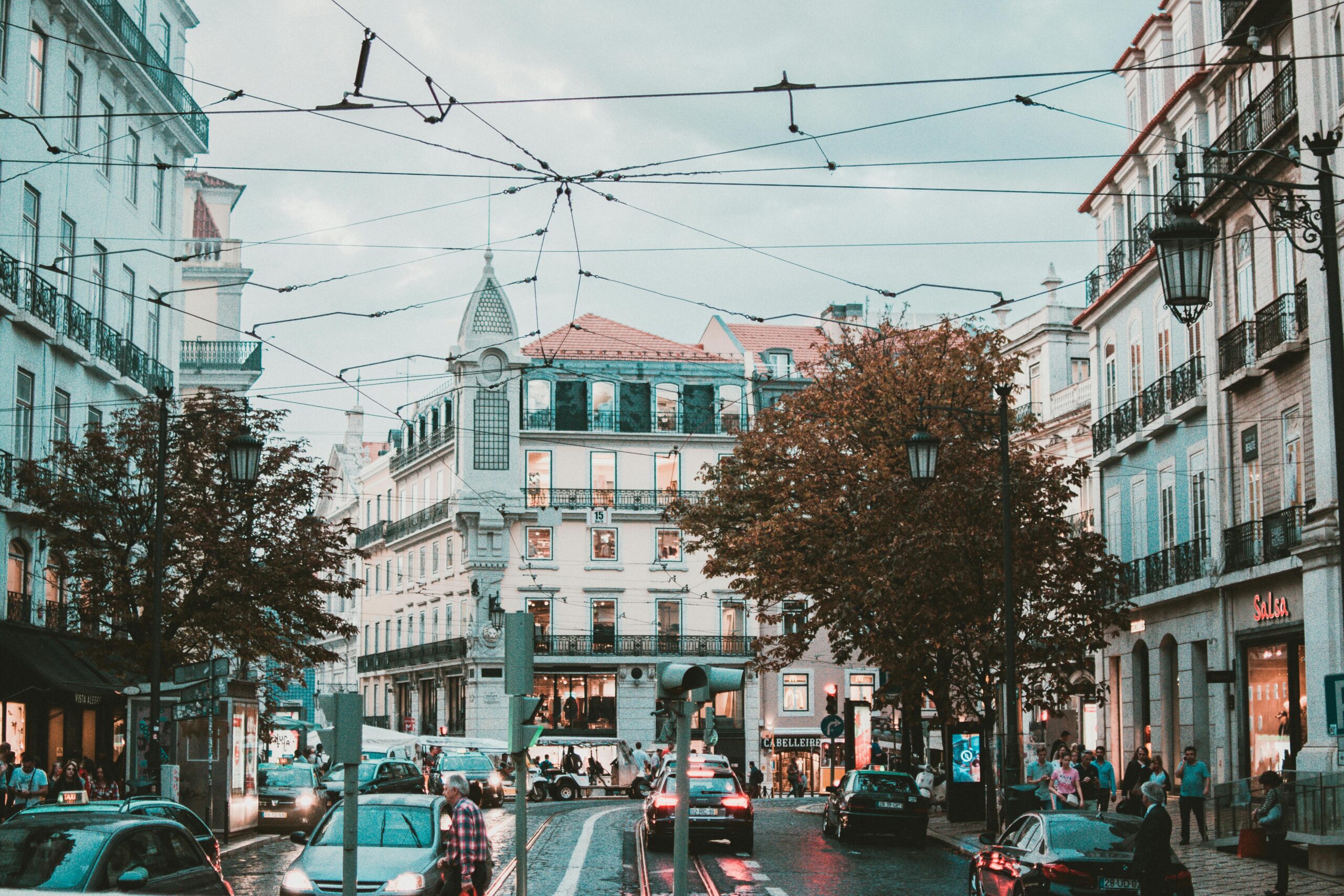 European city street with tram wires, traffic, and historic buildings at dusk in a lively neighbourhood