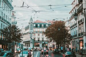 European city street with tram wires, cars, and pedestrians between historic buildings on a cloudy day