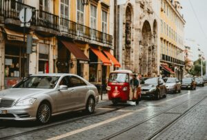 European city street with tram tracks, cars, and a red tuk tuk passing historic buildings and cafes