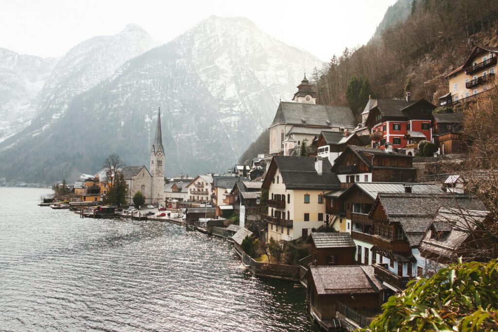 Alpine lakeside village with clustered houses and a tall church spire, backed by misty snow covered mountains.