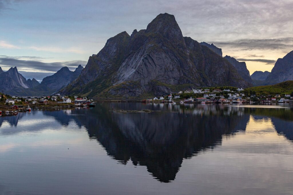 Dusk view of a fjord with a small village and a towering rocky mountain reflected in calm water.