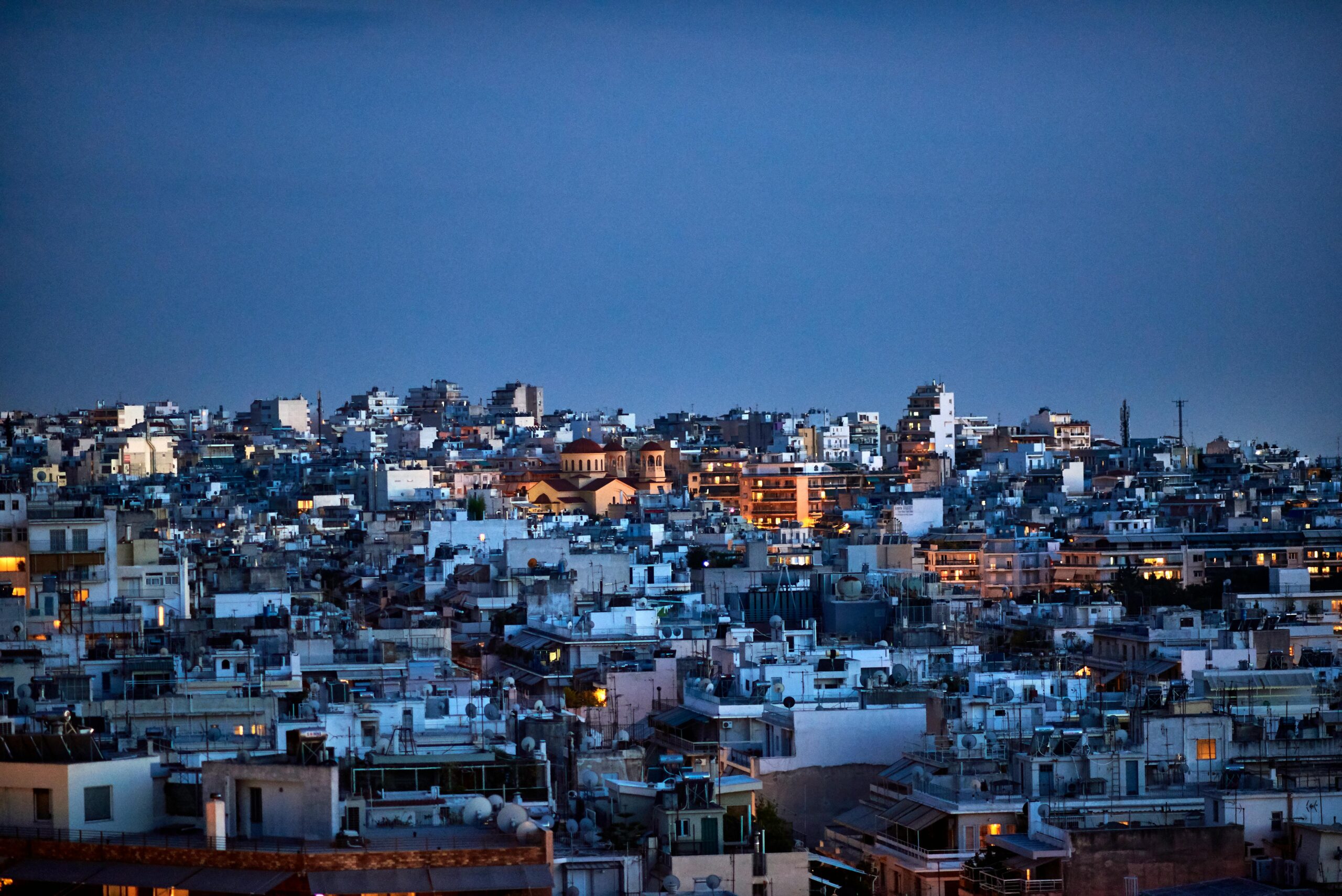 Panoramic view of a dense city at dusk, with rooftops and terraces packed together and warm window lights under a deep blue sky