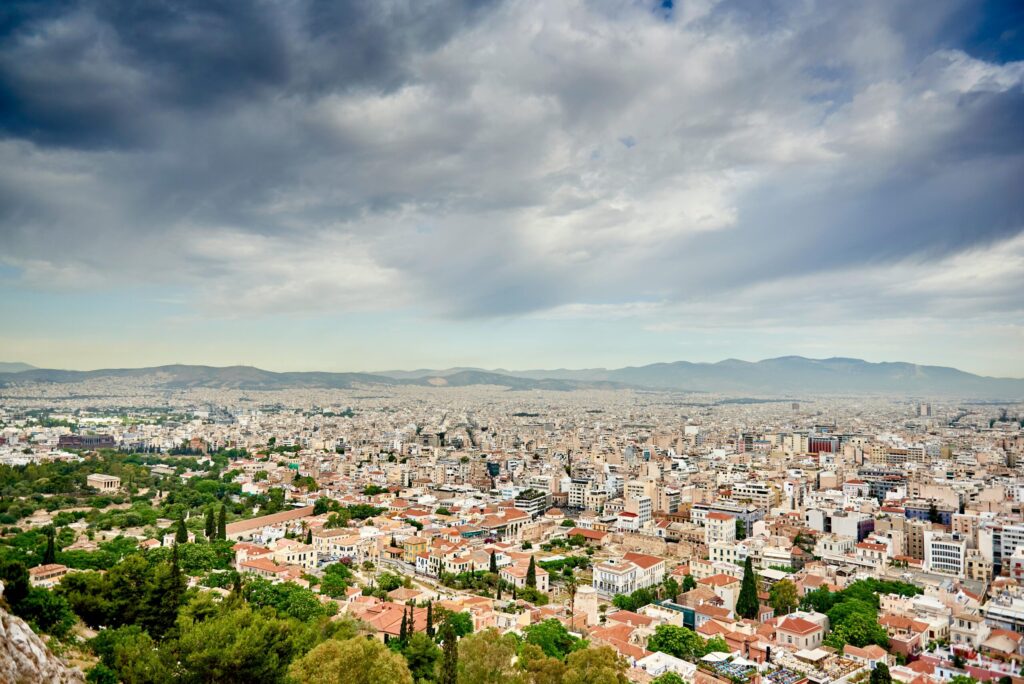 Panoramic view of a sprawling city with terracotta rooftops and green parks in the foreground, mountains on the horizon under dramatic cloudy skies