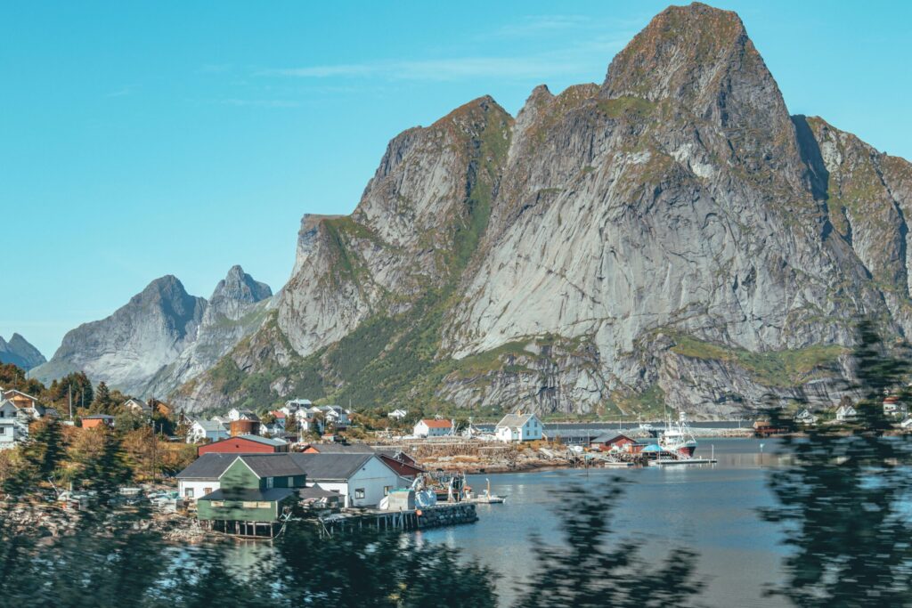 Coastal village beside calm water, with small houses and boats in the foreground and a towering rocky mountain under a clear blue sky.