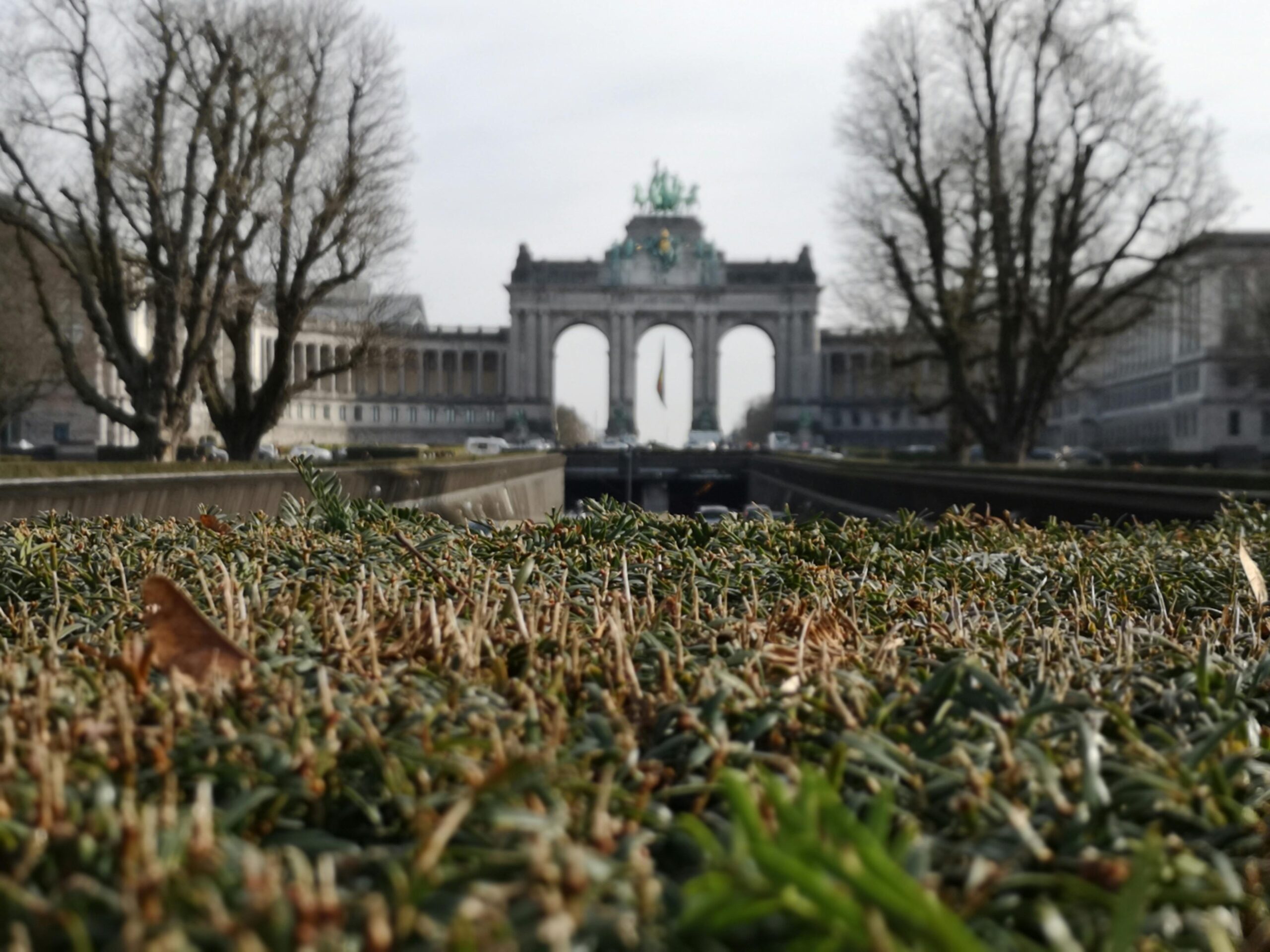 Cinquantenaire Arch in Brussels seen from a low angle over trimmed hedges on a cloudy day, representing moving to Belgium with VANonsite European removals