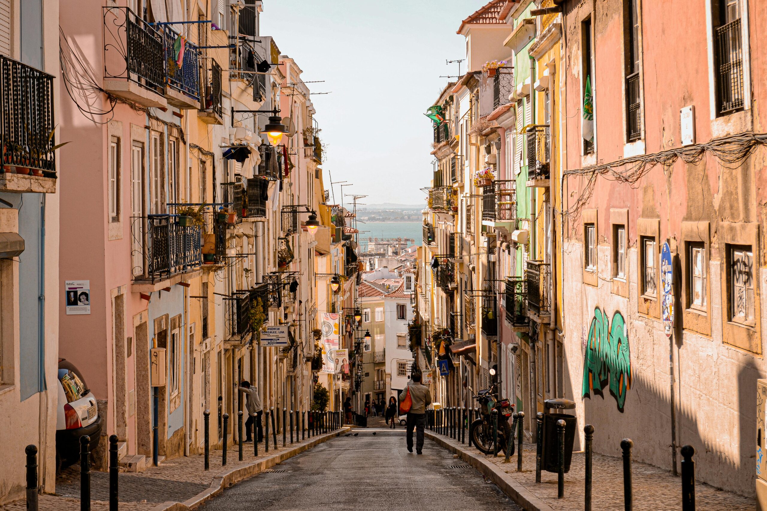Narrow European street lined with colourful apartment buildings and balconies, leading downhill toward the waterfront