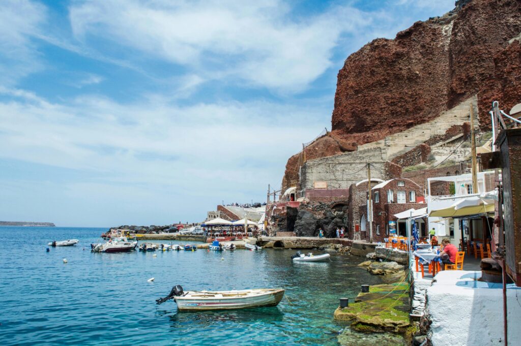 Small boats in a calm harbor beside white waterfront buildings and outdoor cafés, below a steep red cliff