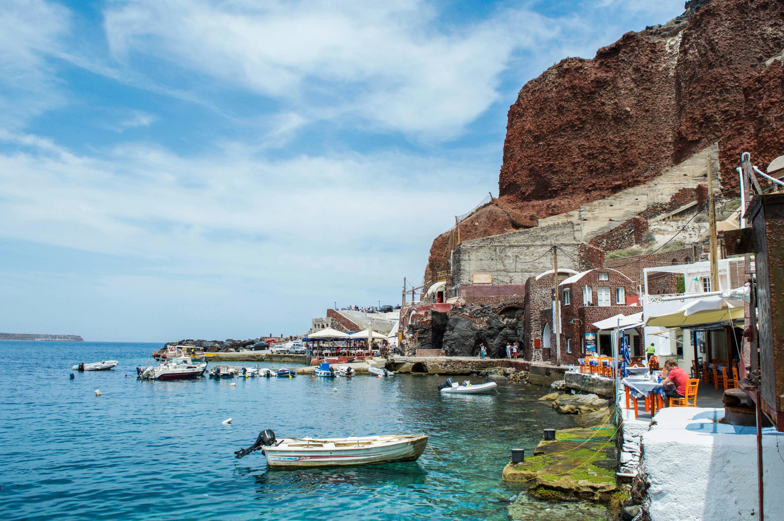 Small boats in a calm harbor beside white waterfront buildings and outdoor cafés, below a steep red cliff
