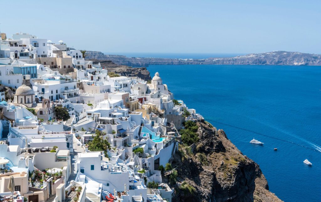 Whitewashed cliffside buildings overlooking a deep blue sea with boats in the distance