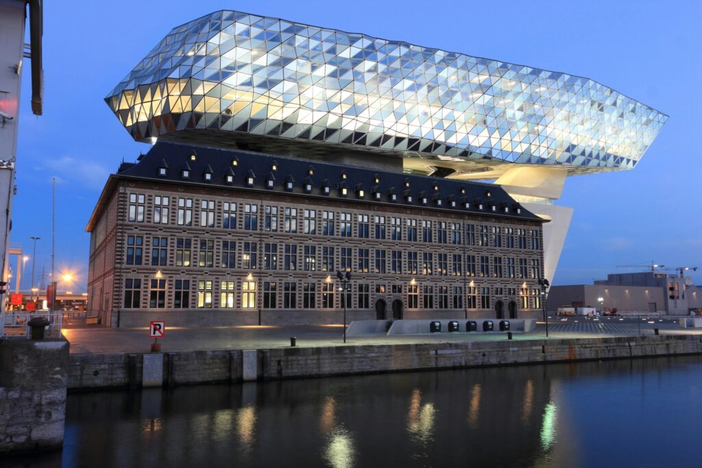 VANonsite moving truck viewpoint on the Port House in Antwerp, a modern glass office above a historic warehouse, symbol of international removals across Belgium and Europe at sunset.