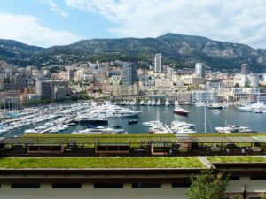 Coastal city marina filled with yachts, dense waterfront skyline, and green mountains under a partly cloudy sky.