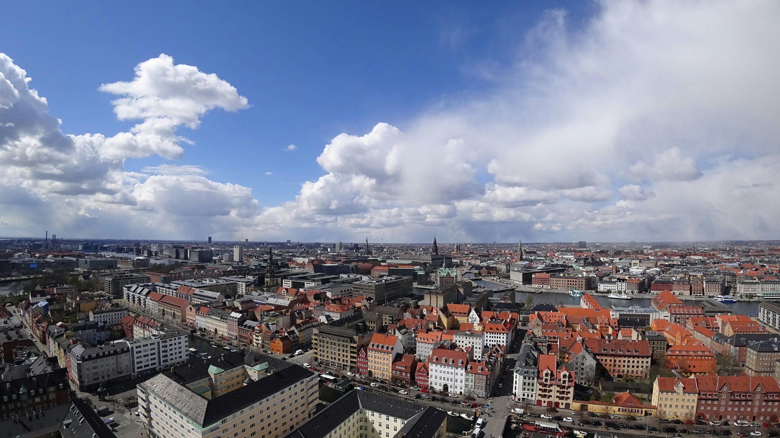 Wide aerial view of a historic city with red rooftops, canals, and landmarks under a bright sky with large clouds.