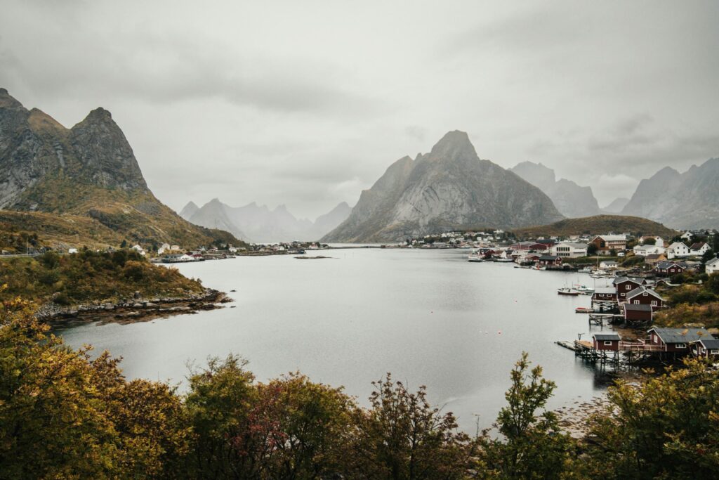 Misty fjord landscape with steep mountains, calm water, and a small village of red and white houses along the shoreline.