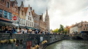 Crowds relaxing by the canal in Bruges, Belgium, with historic brick houses and a stone bridge, showing the lifestyle you can enjoy after moving to Belgium with VANonsite international removals